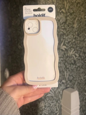 Beige vågigt mobilskal från Holdit - Helt nytt skal, men har 15 PRO så den passar tyvärr inte min mobil….                            Snyggt mobilskal från Holdit i beige med vågig kant och transparent baksida. Passar iPhone 13/14/15/17e/16e. Skalets material är en kombination av mjuk plast och silikon som ger bra grepp och skydd. Perfekt för dig som vill ha ett stilrent men ändå unikt skal.