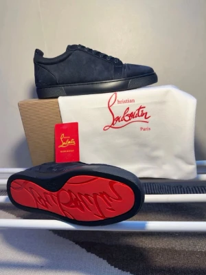 Mörkblåa Louboutin sneakers med röd sula - Snygga Mörkblåa sneakers från Christian Louboutin i mocka med den ikoniska röda sulan. Lågt skaft och klassisk siluett som passar till många stilar. Skorna har svarta snören och diskreta detaljer för en clean look.