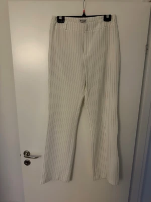 Vita kostymbyxor med svarta ränder - Säljer ett par vita kostymbyxor från Madlady med tunna svarta vertikala ränder. Byxorna har hög midja och raka, långa ben som ger en clean och stilren look. Perfekta för dig som gillar en trendig och klassisk stil.