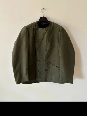 Olivgrön svensk militärjacka med dubbelknäppning - Stilren olivgrön skepparkavaj/armejacka/militärjacka med klassisk dubbelknäppning och två snedställda fickor framtill. Jackan har en rak passform och är tillverkad i ett kraftigt tyg som ger en robust känsla. Perfekt för dig som gillar tidlös och militärinspirerad stil. Insidan är sherpa och utsidan är bomull, 1960s