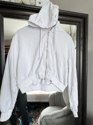 Vit cropped hoodie med dragkedja - Säljer en vit cropped hoodie med dragkedja framtill och huva med dragsko. Hoodien har långa ärmar och bred mudd i nederkant. Materialet är mjukt och känns som bomullsmix, perfekt för en chill och avslappnad stil.