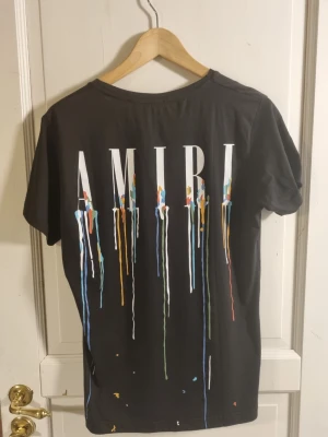Svart AMIRI t-shirt med färgstänk - Snygg svart t-shirt från AMIRI med stort tryck på ryggen och mindre tryck fram. T-shirten har färgstänk i flera nyanser och AMIRI-logga i vitt. Klassisk passform och rund halsringning. Perfekt för dig som gillar streetwear och vill sticka ut.