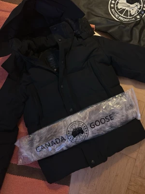 Svart Canada Goose wyndham jacka - Säljer en svart dunjacka från Canada Goose med avtagbar pälskant till huvan. Jackan har hög krage, dragkedja och knappar framtill samt flera praktiska fickor. Perfekt för kalla vinterdagar och riktigt varm tack vare dunfyllningen.