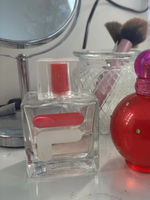 Parfym i fyrkantig glasflaska - Säljer en parfym i en stilren, fyrkantig glasflaska med genomskinligt lock och rosa detaljer. Flaskan har en modern design med en vit och rosa logotyp på framsidan. Perfekt att ha på sminkbordet för en fräsch touch.