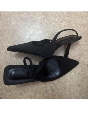 Svarta spetsiga pumps med öppen häl - Stilrena svarta pumps med spetsig tå och öppen häl. Skorna har en smal rem runt hälen och en elegant klack. Ovandelen är i tyg och innersulan är lätt vadderad för extra komfort. Perfekta för dig som gillar en klassisk men trendig look. Använd endast några timmar, säljer på grund av storleksfel 