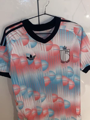 Belgien tröja - Säljer en belgientröja från Adidas med färgglatt mönster i rosa, blått och vitt. Tröjan har svarta detaljer vid krage och ärmslut samt klassiska Adidas-ränder på axlarna. Perfekt för dig som vill sticka ut på träningen!