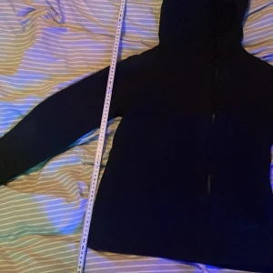 Svart vindjacka från Nike med huva - Snygg svart vindjacka från Nike med hel dragkedja framtill och praktisk huva. Jackan har diskret Nike-logga på bröstet och är tillverkad i ett lätt, vindtätt material. Perfekt för blåsiga dagar och enkel att matcha med olika outfits.