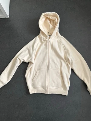Krämvit hoodie med dragkedja - Säljer en krämvit hoodie med dragkedja framtill och stor ficka på magen. Hoodien har huva med dragsko och ribbade muddar vid ärmar och nederkant. Tillverkad i mjukt bomullsmaterial, perfekt för chill dagar.