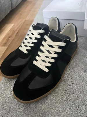 Maison margiela - Stilrena svarta sneakers med vita snören och beige sula. Skorna har en kombination av mocka och slätt läder på ovandelen, samt en klassisk låg silhuett. Perfekta för dig som gillar en clean och tidlös look.