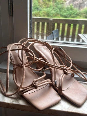 Beige sandaletter med snörning - Säljer ett par trendiga sandaletter från Shein i beige färg med fyrkantig tå och höga klackar. Sandalerna har långa snören som kan lindas runt benet och detaljer i metall på snörändarna. Perfekta för att lyfta din outfit med en modern touch.