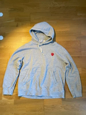 Grå  hoodie Comme des Garçons Play lightly distressed - Säljer en grå hoodie från Comme des Garçons Play med det ikoniska röda hjärtat på bröstet. Hoodien har dragsko i huvan och två fickor framtill. Tillverkad i mjukt bomullsmaterial, perfekt för en avslappnad och stilren look. Är distressed för stil, 100% autentisk.