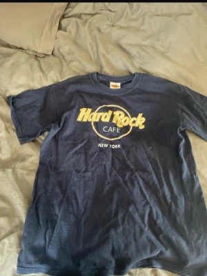 Blå Hard Rock Cafe t-shirt New York - Säljer en klassisk mörkblå Hard Rock Cafe t-shirt med New York-tryck framtill. T-shirten är i 100% bomull och har en normal passform med korta ärmar. Perfekt för dig som gillar ikoniska tryck och vill ha en avslappnad stil.