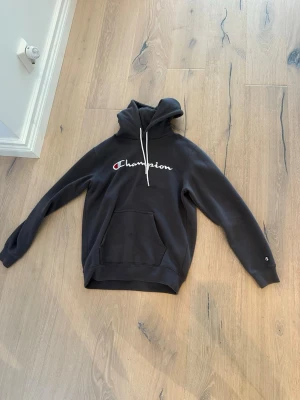 Mörkgrå Champion hoodie med huva - Säljer en mörkgrå hoodie från Champion med stor broderad logga på bröstet och vita dragsnören i huvan. Klassisk känguruficka framtill och ribbade muddar vid ärmar och nederkant. Perfekt för chill dagar eller när du vill ha en sportig look.