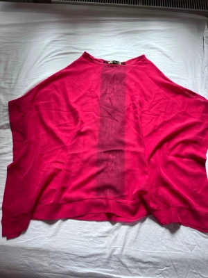 Vid rosa poncho-lik topp med spets och rosetter - Säljer en oversized rosa topp med bred passform. Baksidan har en svart spetsdetalj längs mitten och fyra svarta rosetter. Materialet är tunt och flowy, perfekt för dig som gillar statement-plagg med unika detaljer.