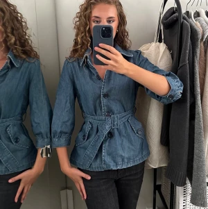 Blå jeansjacka med bälte - Snygg blå jeansjacka med markerad midja och tillhörande bälte. Jackan har trekvartsärmar, stora knappar framtill och fickor med lock. Perfekt för dig som vill ha en trendig och unik jeansjacka med cool siluett.