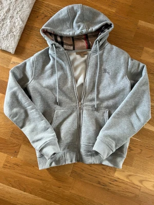 Grå zip hoodie med huva - Snygg grå hoodie med dragkedja och huva. Klassisk passform med ribbade muddar och fickor framtill. Insidan av huvan har ett rutigt mönster som ger en extra detalj. Perfekt för en avslappnad stil.