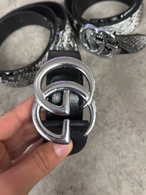 Svart bälte med silverfärgat spänne - Snyggt svart bälte i skinn med ett stort, rundat silverfärgat spänne framtill. Bältet har en klassisk design och passar perfekt till jeans eller byxor. Insidan är brun och utsidan är slät och stilren.