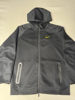 Nike Nocta - Snygg mörkgrå hoodie från Nike med hel dragkedja framtill och två fickor med dragkedja. Liten gul Nike-logga på bröstet och diskreta gula initialer samt symbol på ryggen. Tillverkad i ett mjukt, sportigt material och har huva för extra stilpoäng.