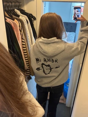 Grå hoodie med tryck 'Mi Amor' - Snygg grå hoodie med svart tryck på ryggen där det står 'Mi Amor' och ett hjärta genomborrat av en pil. Klassisk passform med huva och ribbade muddar. Perfekt för dig som vill ha en unik och trendig stil.