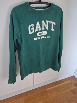 Gant Sweatshirt - Mysig grön sweatshirt från GANT med vit text 'GANT 1949 NEW HAVEN' på bröstet. Tröjan har rund halsringning, långa ärmar och ribbade muddar. Perfekt för en avslappnad och sportig stil.