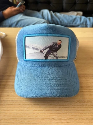 Blå Maggiore truckerkeps - Snygg blå truckerkeps från Maggiore med mesh på sidorna och baksidan. Framsidan har en patch med bildmotiv och logga. Skärmen är i mockaliknande material och kepsen har justerbar rem bak. Perfekt för en avslappnad streetwear-look.