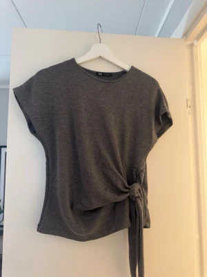 Grå topp med knyt från Zara - Säljer en enkel och snygg grå topp från Zara med korta ärmar och knyt i sidan. Toppen har rund halsringning och är gjord i mjukt material som känns skönt mot huden. Perfekt till jeans eller kjol för en chill look.