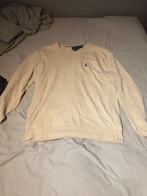 Stickad tröja Ralph lauren - Stilren beige tröja med rund halsringning och diskret broderad logga på bröstet. Långärmad modell med ribbade muddar vid ärmslut och nederkant. Perfekt för en clean och enkel look.