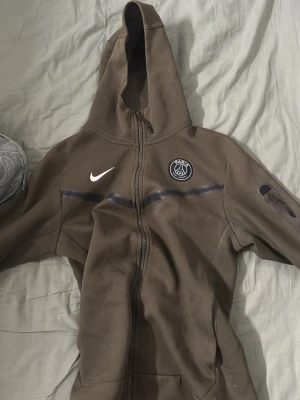 Brun Nike PSG hoodie med dragkedja - Säljer en brun hoodie från Nike med PSG-logga på bröstet och hel dragkedja framtill. Hoodien har huva, ficka med dragkedja på ärmen och diskreta svarta detaljer över bröstet. Perfekt för dig som gillar sportig stil och vill representera Paris Saint-Germain. Den e äkta kom  med bud säljs ej längre i butik 