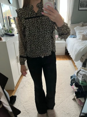 Leopardmönstrad blus med volang - Snygg leopardmönstrad blus med volangdetaljer vid axlarna och svart band över bröstet. Blusen har lång ärm och är gjord i ett tunt, lätt material som ger en luftig känsla. Perfekt för dig som vill sticka ut med ett trendigt djurmönster.