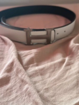 Vitt bälte med silverfärgat H-spänne - Snyggt vitt bälte med ett stort, silverfärgat H-format spänne. Bältet har en stilren design och passar perfekt till jeans eller byxor för att lyfta din outfit. Materialet ser ut att vara skinn och insidan är mörkare för en cool kontrast.