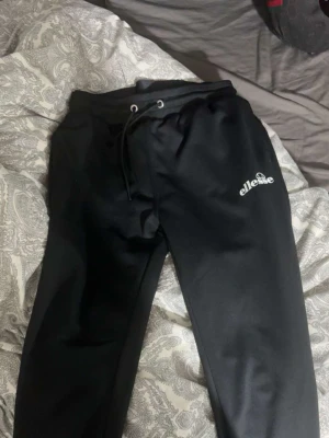 Ellesse byxor - Svarta mjukisbyxor från Ellesse med dragsko i midjan och logga på vänster lår. Byxorna har en avslappnad passform och är tillverkade i ett mjukt material som känns skönt mot huden. Perfekta för chill dagar eller när du vill ha det bekvämt.
