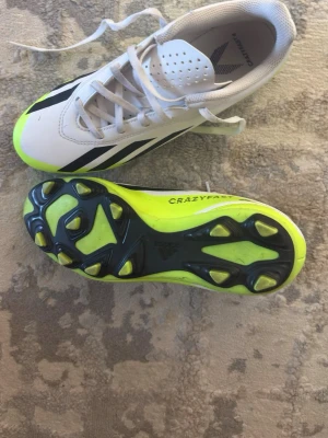 Vita och neongula fotbollsskor - Säljer ett par snygga fotbollsskor i vitt med svarta detaljer och neongul sula. Skorna har klassisk snörning och greppvänliga dobbar för bra fäste på planen. Perfekta för dig som vill sticka ut på matchen! Str 38/39