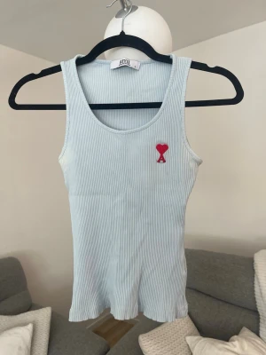 Ljusblå ribbad linnetopp från Ami - Säljer en ljusblå ribbad linnetopp från Ami Alexandre Mattiussi med broderad röd logga på bröstet. Toppen är ärmlös och har en tight passform, perfekt för en stilren och enkel look.