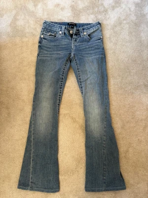 Blåa true religion jeans - Säljer ett par blå true religion  jeans  i storlek W23 med  snygga detaljer på bakfickorna. Jeansen har låg midja, fem fickor och dekorativa knappar bak. Klassisk tvättad denim som passar till det mesta. Materialet är bomullsdenim. Pris kan diskuteras vid snabbaffär💕Byxorna är uppsydda därför är innerbenslängden 72cm