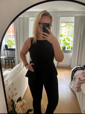 Svart ärmlös topp med rosett - Snygg svart topp med smala axelband och peplumformad nederdel. Toppen är ärmlös och har en öppen rygg med en stor rosett som knyts baktill. Perfekt för dig som vill ha en stilren men ändå unik look.