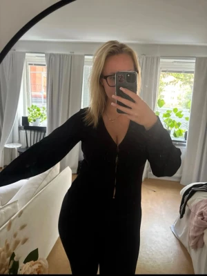 Svart v-ringad topp med hakes stängning  - Snygg svart topp med v-ringning och hakes stängning framtill. Långärmad modell i mjukt material som sitter figurnära. Perfekt att styla med jeans eller kjol för en clean look.