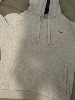 Grå hoodie från Lacoste med huva - Snygg grå hoodie från Lacoste med klassisk krokodillogo på bröstet. Hoodien har huva med dragsko och en stor magficka framtill. Tillverkad i mjukt sweatshirtmaterial som är perfekt för chill dagar.