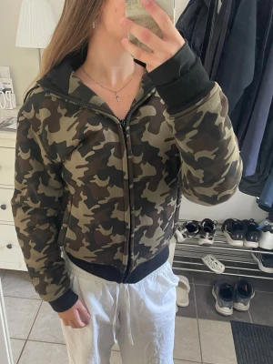 Camouflage bomberjacka med huva - Säljer en snygg bomberjacka i klassiskt camouflage-mönster i gröna och bruna toner. Jackan har svart huva, muddar vid ärmslut och midja samt dragkedja framtill. Perfekt för dig som vill ha en streetwear-inspirerad look. Finns skydd i för axlar, och underarmarna. Kostar 2400 kr ny. Använt nån gpng, gelt ny i skicket