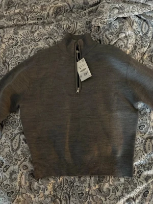 Grå halvzip tröja med hög krage - Snygg grå tröja med halv dragkedja och hög krage. Tröjan har ribbade muddar vid ärmslut och nederkant, vilket ger en clean och modern look. Perfekt till jeans eller  för en avslappnad stil.