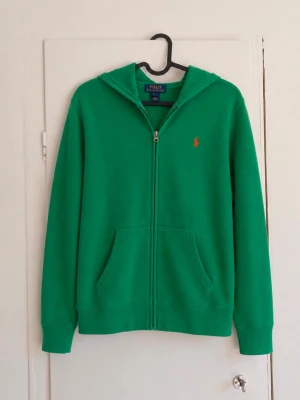 Polo Ralph Lauren zip hoodie grön – L (14–16) 🐎💚 - Snygg grön hoodie från Polo Ralph Lauren med orange broderad logga på bröstet. Klassisk modell med hel dragkedja, luva och fickor framtill. 🔹 Storlek: L (14–16 år) men passar ca xs-s! 🔹 Färg: Grön 🔹 Orange broderad Polo-logo 🔹 Hel dragkedja 🔹 Luva och fickor framtill 🔹 Fint begagnat skick men den är nopprig!😁 Superfin färg och en klassisk Ralph Lauren-modell som passar perfekt till vardags. Hör gärna av dig vid frågor eller om du vill ha mått! 💌