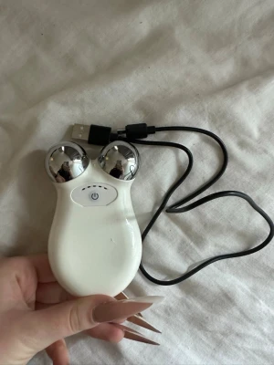 Vitt ansiktsmassageverktyg microströmsapparat med USB - Smidigt ansiktsmassageverktyg i vitt med två silverfärgade kulor och USB-sladd. Perfekt för att stimulera ansiktsmuskler och ge huden en fräsch känsla. Enkel att använda med en knapp på framsidan och laddas via USB.