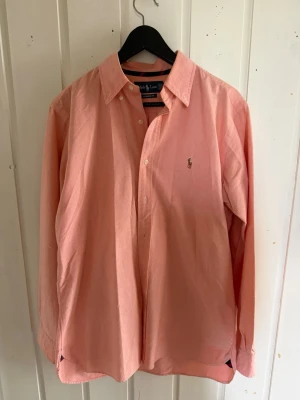 Skjorta från Ralph Lauren - Säljer en klassisk rosa skjorta från Ralph Lauren i modellen Classic Fit. Skjortan har långa ärmar, knappar framtill och den ikoniska loggan broderad på bröstet. Perfekt för dig som vill ha en stilren och tidlös look.