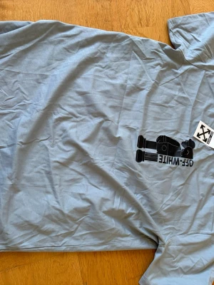 Grå Off-White t-shirt med tryck - Snygg grå t-shirt från Off-White med svart grafiskt tryck framtill och klassisk rund hals. T-shirten har korta ärmar och är gjord i mjuk bomull. Perfekt för dig som gillar streetwear och vill sticka ut med en unik design.
