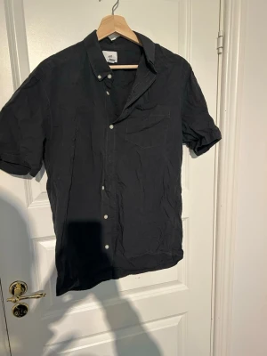 Mörkgrå kortärmad skjorta H&M - Säljer en mörkgrå kortärmad skjorta från H&M i regular fit. Skjortan har klassisk krage med knappar, bröstficka och vita knappar framtill. Perfekt till sommaren och enkel att matcha med olika outfits.