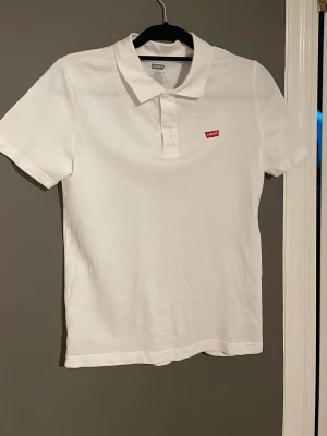 Vit pikétröja från Levi's - Klassisk vit pikétröja från Levi's med korta ärmar och krage. Tröjan har knappar framtill och den ikoniska röda Levi's-loggan på bröstet. Tillverkad i mjuk bomull för en bekväm känsla.