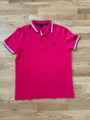 Rosa pikétröja från BOSS - Snygg rosa pikétröja från BOSS med klassisk krage och knäppning framtill. Tröjan har vita och svarta detaljer på krage och ärmslut samt korta ärmar. Perfekt för en stilren och sportig look.