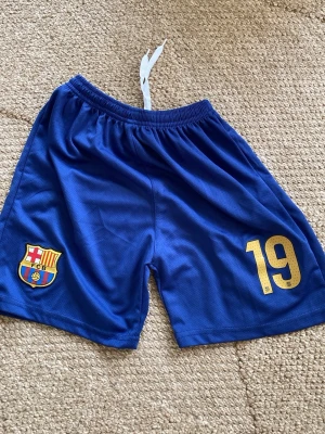 Blå FC Barcelona fotbollsshorts - Säljer ett par blå FC Barcelona shorts med klubbmärke och nummer 19 i gult på ena benet. Shortsen har resårmidja med snörning och är gjorda i ett lätt och luftigt syntetmaterial, perfekta för fotboll eller träning.