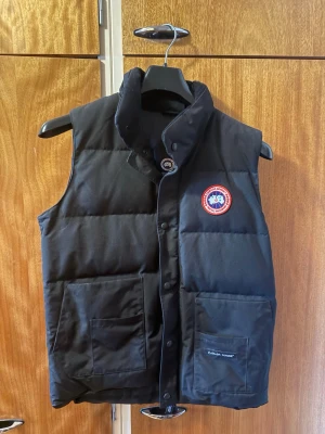 Svart dunväst från Canada Goose - Snygg svart dunväst från Canada Goose med hög krage och två stora fickor framtill. Västen har det klassiska runda Canada Goose-märket på bröstet och stängs med tryckknappar. Perfekt för lager-på-lager och håller dig varm under kyliga dagar.