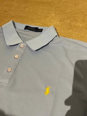 Ljusblå pikétröja från Polo Ralph Lauren - Snygg ljusblå pikétröja från Polo Ralph Lauren med klassisk krage, tre knappar och gul broderad logga på bröstet. Perfekt till jeans eller shorts för en clean och stilren look.