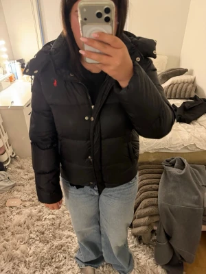 Polo ralph lauren jacka - Säljer en svart pufferjacka med huva och dragkedja samt knappar framtill. Jackan har en röd logga broderad på bröstet och en klassisk quiltad design. Perfekt för kalla vinterdagar och riktigt skön att bära.